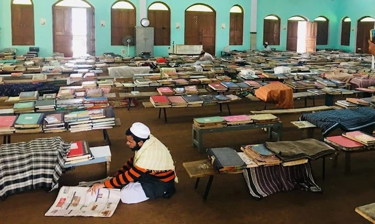 Person reading newspaper in room with rows of beds and books.